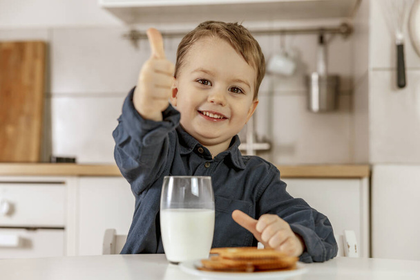 Egy kisfiú ül a konyhában és tejet iszik. Friss tej üvegben, tejtermék egészséges ital. Egészségügyi ellátás, kalciumforrás, laktóz. Hangulatos és modern belső tér. Óvodáskorú gyermek alkalmi ruházattal. - Fotó, kép