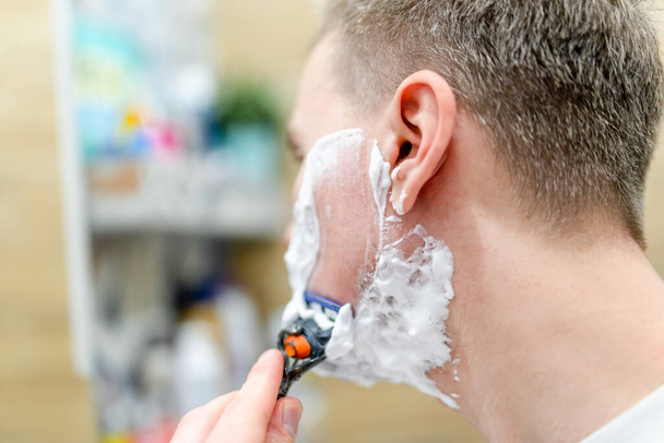 Молодой человек бреется в ванной с зеркалом, используя пену для бритья. Селективный фокус - Фото, изображение