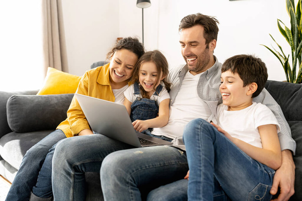 Cheerful happy family of four using laptop sitting on the sofa in cozy apartment - Foto, Imagem