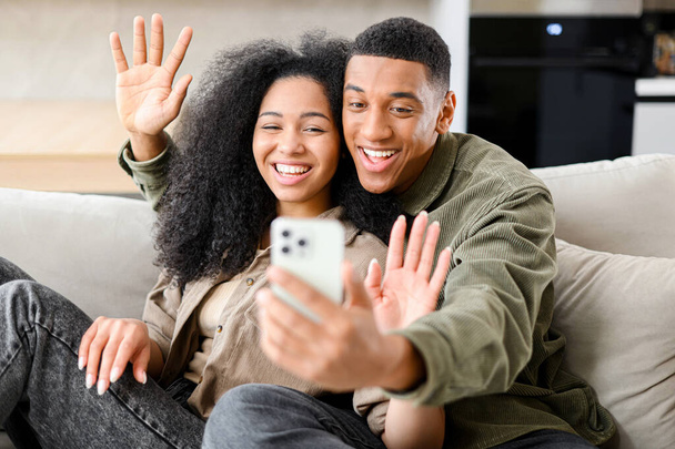 携帯電話でビデオ通話を介してオンラインウェブカメラチャットを持つ幸せな陽気な若い多人種のカップルのフロントビュー,居心地の良いアパートに一緒に座っている間 - 写真・画像