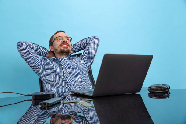 Portrait of happy looking man, computer support specialist, programmer sitting in front of laptop isolated over blue background - Фото, зображення