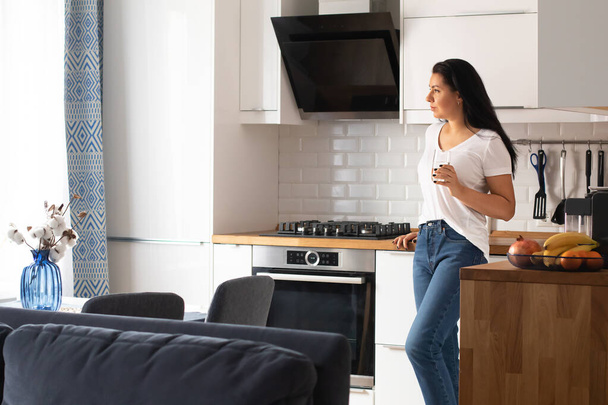 Una giovane donna bruna in jeans e una maglietta bianca beve acqua in cucina - Foto, immagini