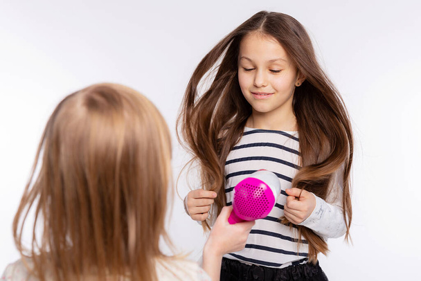 Két gyönyörű 6-8 éves lány játszik a hajszárítóval. Lányok játékos időt töltenek együtt, és szárítás haj felett fehér háttér - Fotó, kép