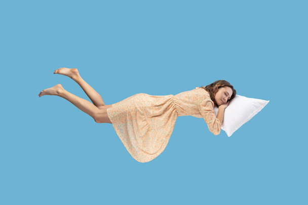 Relaxed girl in yellow dress levitating in mid-air, sleeping on stomach lying comfortable cozy on pillow, keeping eyes closed, watching peaceful dream. indoor studio shot isolated on blue - Foto, immagini