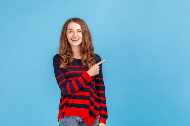 Positive smiling woman wearing striped casual style sweater, pointing aside, showing copy space for commercial text, blank wall with idea presentation. Indoor studio shot isolated on blue background. - Фото, изображение