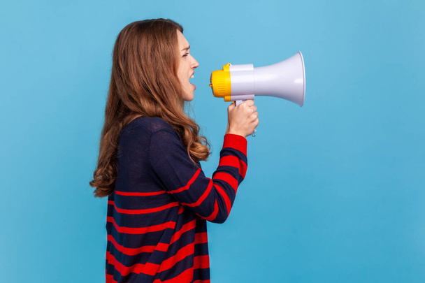 Side view of woman wearing striped casual style sweater, standing screaming in megaphone, announcing important information or protesting. Indoor studio shot isolated on blue background. - Valokuva, kuva