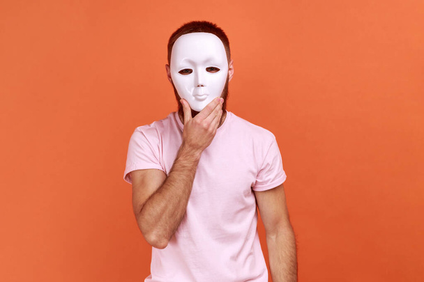 Portrait of unknown bearded man covering her face with white mask, hiding her real personality, anonymity, wearing pink T-shirt. Indoor studio shot isolated on orange background. - Fotografie, Obrázek