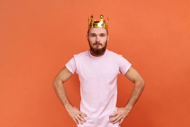 Porträt eines bärtigen Mannes, der egoistisch in die Kamera blickt, mit Krone auf dem Kopf posiert, sich als König ausgibt, privilegierten Status genießt und rosa T-Shirt trägt. Indoor Studio isoliert auf orangefarbenem Hintergrund aufgenommen. - Foto, Bild