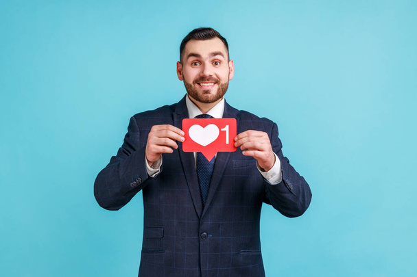 Satisfied man with beard wearing official style suit holding heart icon, recommending to follow and share blog with interesting content. Indoor studio shot isolated on blue background. - Photo, image