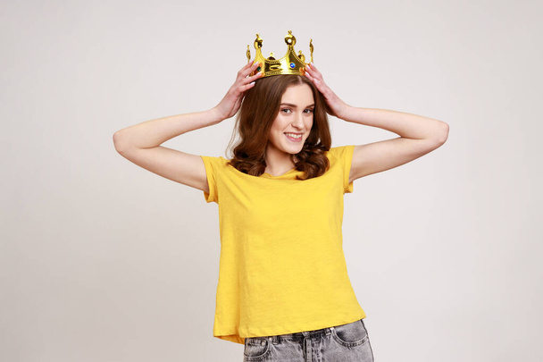 I am queen! Portrait of happy curly-haired teenager girl putting crown on head and smiling, self confidence, self-motivation and dreams to be best. Indoor studio shot isolated on gray background. - Photo, image