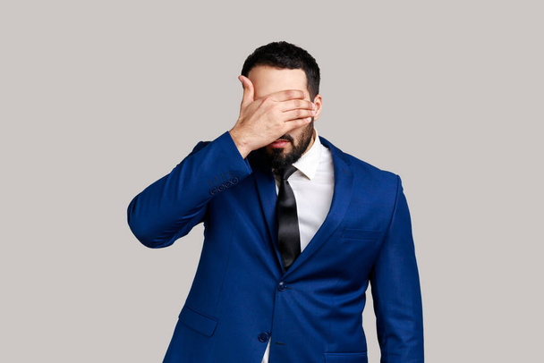 Bearded man closing eyes with hand, dont want to see that, ignoring problems, hiding from stressful situations, wearing official style suit. Indoor studio shot isolated on gray background. - 写真・画像