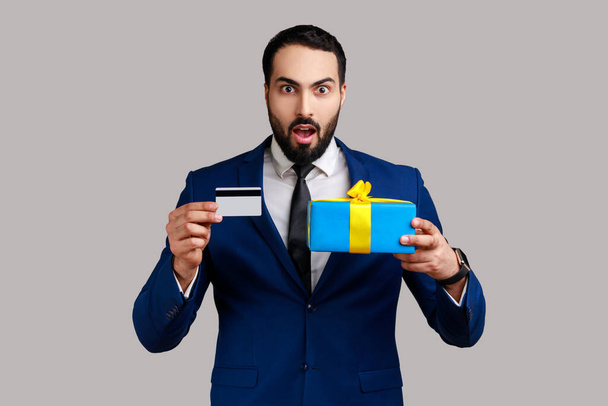 Portrait of shocked astonished bearded man showing credit card and gift box, looking with amazement, banking, wearing official style suit. Indoor studio shot isolated on gray background. - Foto, afbeelding