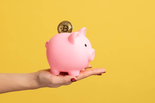Close up side view portrait of woman hand holding piggy bank with golden coin, savings of money. Indoor studio shot isolated on yellow background. - Fotó, kép
