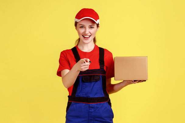 Portrét usměvavé optimistické doručovatelky stojící s kartonovou krabičkou, balíček pro klienta, ukazující na kameru, v montérkách a červené čepici. Vnitřní studio záběr izolované na žlutém pozadí. - Fotografie, Obrázek