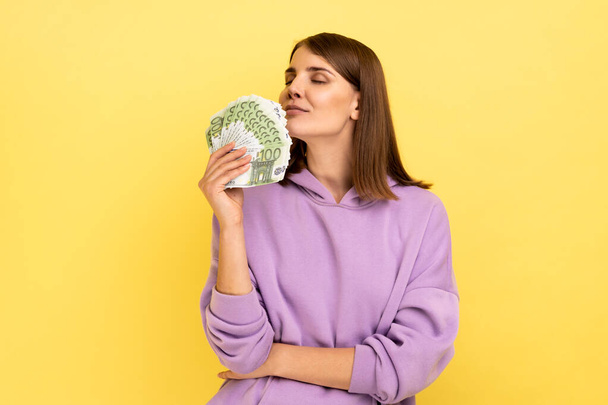 Portrait of attractive young rich woman holding big fan of money, smelling euro banknotes with pleasure, wearing purple hoodie. Indoor studio shot isolated on yellow background. - Valokuva, kuva