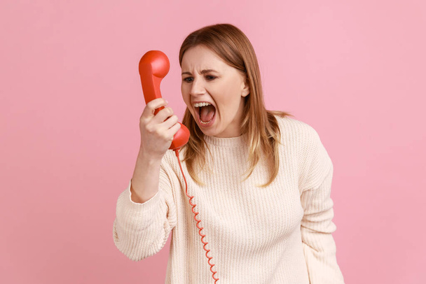 Portrait of angry upset blond woman screaming into red handset, having unpleasant conversation, expressing aggression, wearing white sweater. Indoor studio shot isolated on pink background. - Foto, afbeelding