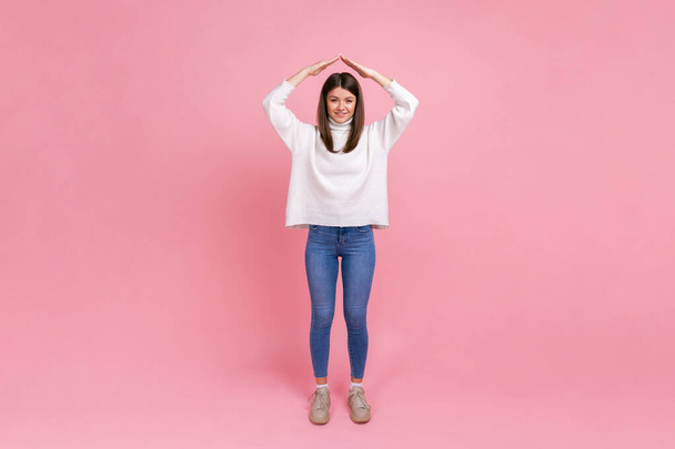 Full length portrait of attractive woman making roof of hands above head, feeling herself in safety, wearing white casual style sweater. Indoor studio shot isolated on pink background. - Foto, Bild