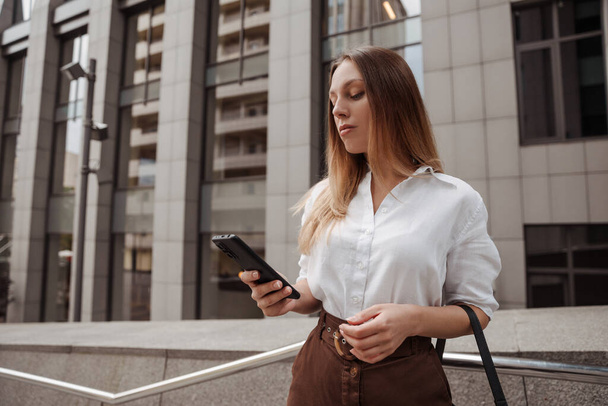низкий угол зрения вдумчивой молодой предпринимательницы, смотрящей на смартфон на городской улице - Фото, изображение