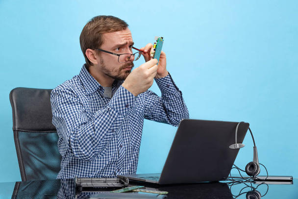Portrait of man in glasses and checkered shirt attentively looking at motherboard isolated over blue background - Fotó, kép