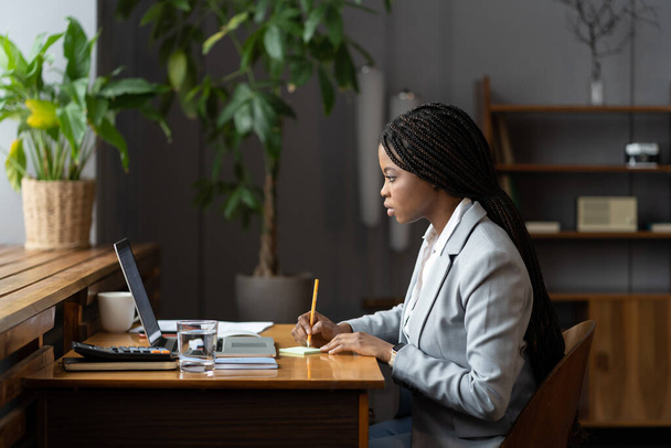 若いアフロビジネス女性研修生がノートパソコンでオンラインで仕事をするオフィスのデスクでビジネスレポートのためのメモを作る - 写真・画像