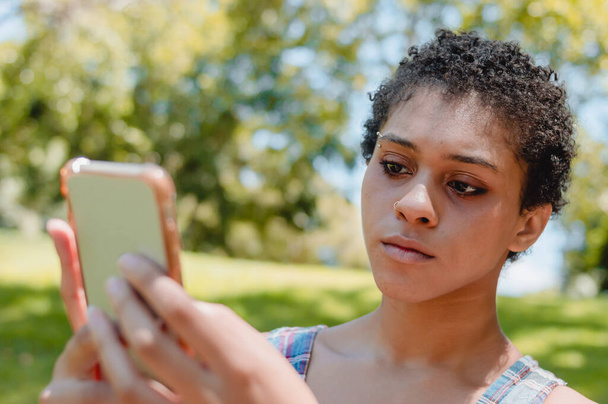 若いラテン系のブルネットの女性は、彼女の携帯電話を見てメッセージを送信し、インターネットを使用して、木や自然とコピースペースを持つ公園で. - 写真・画像
