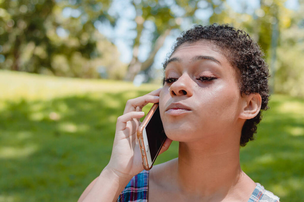brunette argentinian hispanic latin female teenager with short hair attentively listening conversing on a phone call in a park with trees behind her and with copy space - Photo, Image