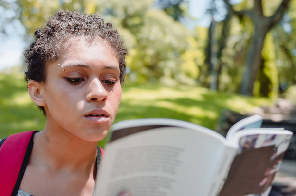 lähikuva muotokuva keskittynyt nuori latino argentiinalainen brunette lyhyet hiukset tyttö lukee kirjaa puistossa, jossa puita taustalla ja kopioi tilaa, kirja päivä ja koulun käsite. - Valokuva, kuva