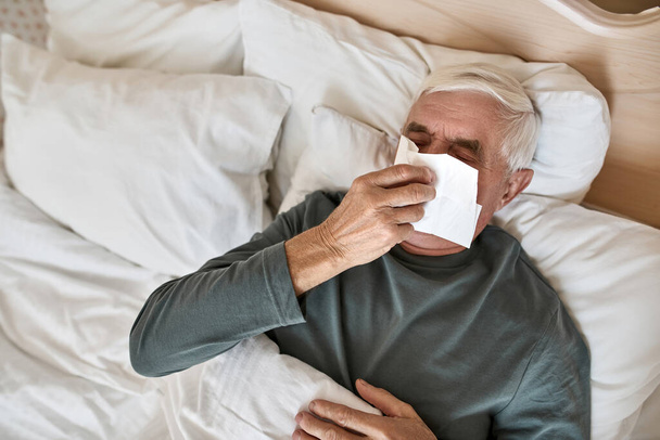 Elderly man blowing runny nose with napkin on bed - Foto, immagini