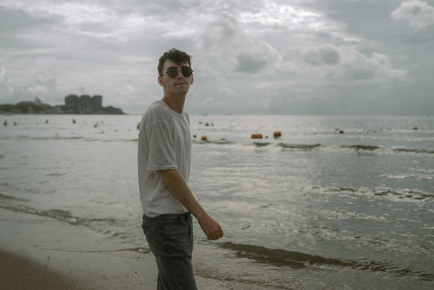 a young guy in sunglasses near the sea looks at the camera, against the backdrop of the city, pursing his lips - Foto, imagen