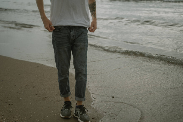 piernas de un hombre caminando en una playa de arena en zapatillas de deporte y jeans - Foto, Imagen