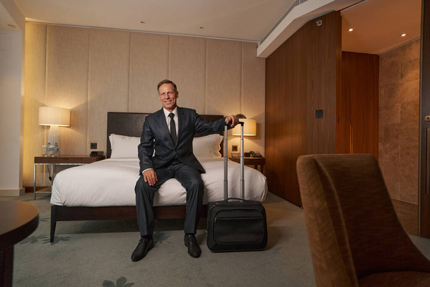 Φιλικός bussinesman χαμογελά ενώ κάθεται στο κρεβάτι του ξενοδοχείου - Φωτογραφία, εικόνα