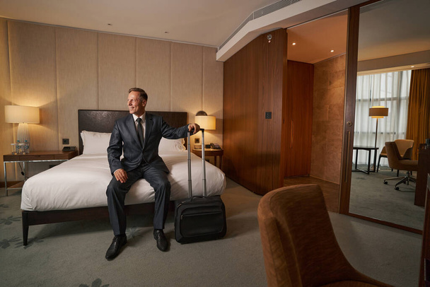 Ενθουσιαστικός επιχειρηματίας κάθεται στο κρεβάτι και ελέγχει τη σουίτα του ξενοδοχείου του - Φωτογραφία, εικόνα