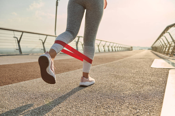 Κοντινό πλάνο. Οπίσθια όψη των ποδιών μιας αθλήτριας, αθλήτριας με γκρίζα πόδια που κάνει προπόνηση σωματικού βάρους, γυμνάζεται με λάστιχο αντοχής σε γέφυρα της πόλης την αυγή - Φωτογραφία, εικόνα