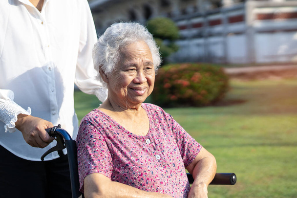 Φροντιστής βοήθεια και φροντίδα ασιατική ανώτερος ή ηλικιωμένη ηλικιωμένη γυναίκα ασθενή κάθεται σε αναπηρική καρέκλα στο πάρκο, υγιή ισχυρή ιατρική έννοια. - Φωτογραφία, εικόνα