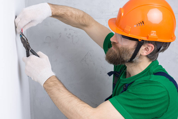 Ein Elektriker installiert Steckdosen in der Wohnung. Ein Mann mit orangefarbenem Helm und Overall macht Elektrik im Haus. - Foto, Bild