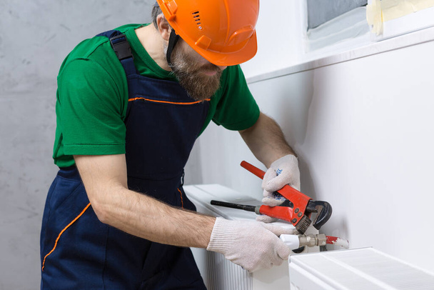 Ein männlicher Klempner installiert einen Heizkörper in der Heizungsanlage einer Wohnung. Kerl in Overalls und einem Gasschlüssel. - Foto, Bild