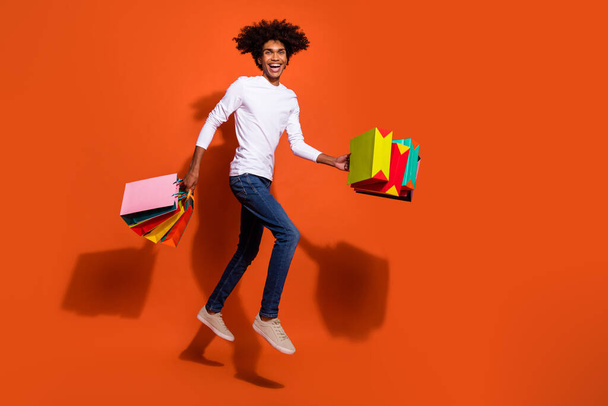 Photo of excited funky guy dressed white shirt holding shoppers jumping high empty space isolated orange color background - Фото, изображение
