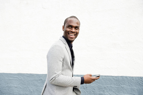 Boczny portret uśmiechniętego afro-amerykańskiego biznesmena chodzącego z komórką przy ścianie - Zdjęcie, obraz