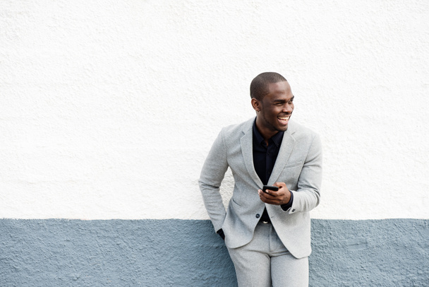 Portret van een gelukkige Afro-Amerikaanse zakenman leunend tegen de muur met een mobiele telefoon - Foto, afbeelding