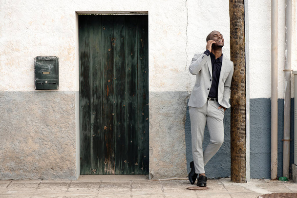 Ganzkörperporträt eines lachenden afrikanisch-amerikanischen Geschäftsmannes, der an der Wand steht und mit dem Handy spricht - Foto, Bild
