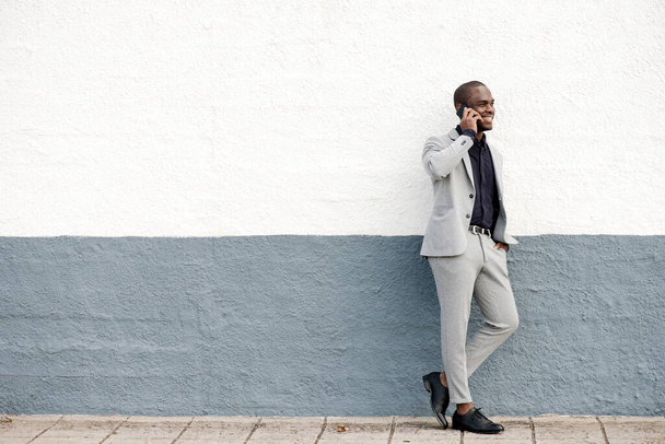 Retrato de cuerpo completo del hombre de negocios afroamericano sonriente parado contra la pared hablando con el teléfono - Foto, Imagen
