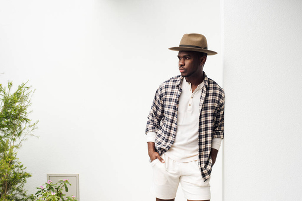 Portrét pohledného afrického Američana opírajícího se o bílou zeď s košilí a kloboukem - Fotografie, Obrázek
