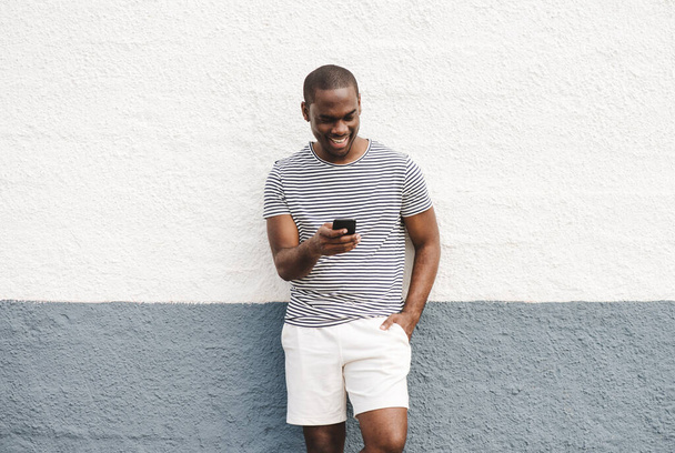 Retrato sonriente afroamericano hombre en pantalones cortos y camiseta mirando el teléfono celular  - Foto, Imagen