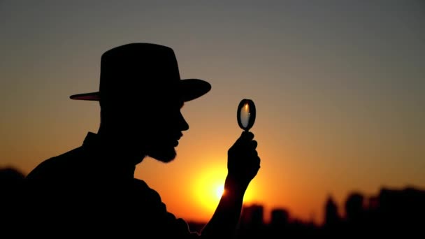 Sylwetka mężczyzna patrząc za pomocą szkła powiększającego o zachodzie słońca - Materiał filmowy, wideo