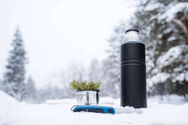 A black matte thermos, a knife and a mug with a spruce twig stand on a snow-covered stump.Warm tea in the winter frost.Minimalistic subject shot. High quality photo - Photo, image