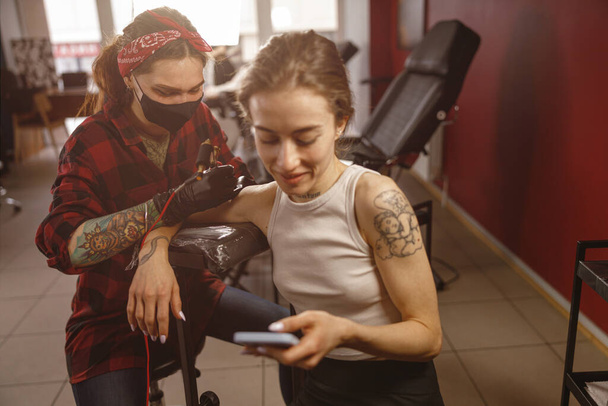 Relaxed woman client using phone while sitting in tattoo salon - 写真・画像