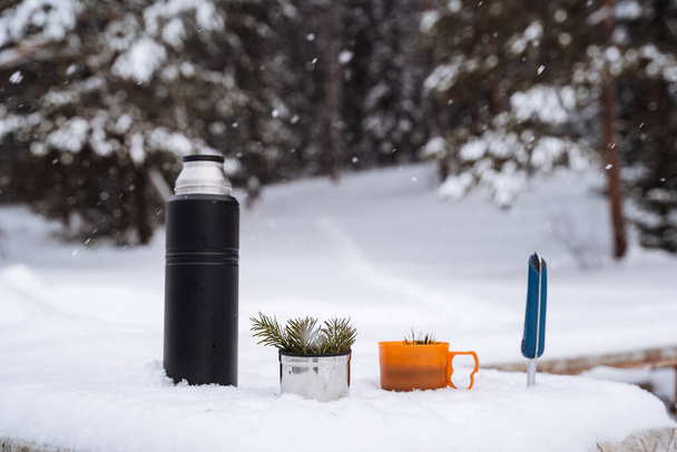 A black thermos and mugs with el's sprig in the snow. A hiking knife will be stuck in the snow. Winter still life in the snow. High quality photo - Photo, image