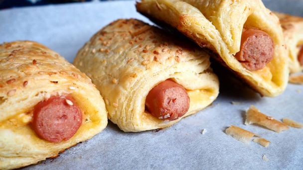 Sausages in the dough on a baking sheet with baking paper. Delicious fresh pies from the oven. Yeast dough culinary products with milk sausages. Sprinkling with sesame seeds - Фото, изображение