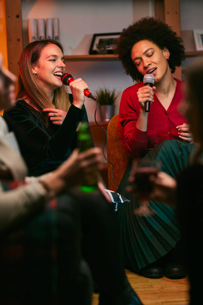 Szczęśliwych wielokulturowych przyjaciół siedzących w domu, śmiejących się, śpiewających i bawiących się w noc karaoke. - Zdjęcie, obraz