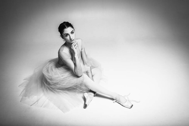 Tired pretty, emotional ballerina sitting on light background background, place for inscription. Black and white photo - Фото, зображення